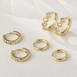 May include: A selection of gold-coloured hoop earrings. Some hoops are decorated with small, square-shaped clear stones. The earrings vary in size and design, with some featuring a continuous row of stones and others with a few larger stones.