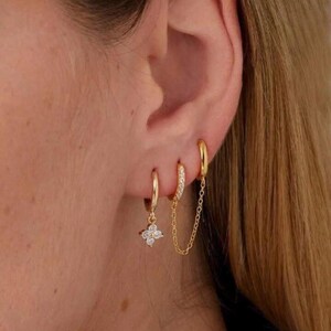 May include: A set of gold-coloured hoop earrings. One earring has a small, dangling flower design with clear stones. Another has a chain connecting it to a second hoop. The third hoop is a simple, solid gold colour. The earrings are worn on an ear.