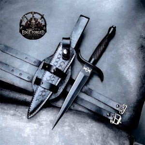 May include: A black dagger with a textured handle and a white geometric symbol on the blade, accompanied by a black leather sheath featuring decorative stitching and two buckled straps. The logo for Edge Forged Knives is visible in the corner.