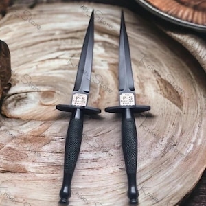 May include: Two black F-S Fighting Knives with sharp, pointed blades and textured handles. The knives have a crossguard and a small rectangular emblem with text. They are displayed on a wooden surface.
