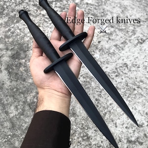 Puede incluir: Dos dagas negras con mangos texturizados y guardas. Las hojas son afiladas y puntiagudas. El texto "Edge Forged knives" es visible en la imagen.