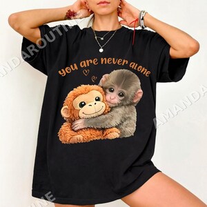 May include: Black t-shirt with a graphic of two monkeys embracing, accompanied by the orange text "you are never alone". The monkeys are depicted in brown and grey, with smiling expressions and two small hearts.