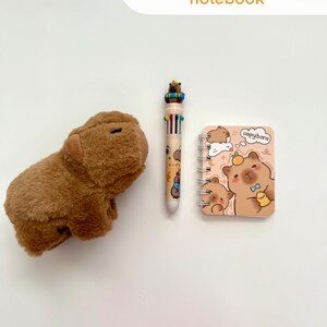Puede incluir: Un capibara de peluche marr&oacute;n, un bol&iacute;grafo multicolor con un dise&ntilde;o de capibara y un peque&ntilde;o cuaderno con espiral con una ilustraci&oacute;n de capibara. El texto "Capybara, pen & notebook" est&aacute; en la parte superior.