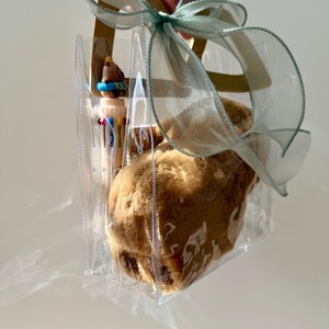 Puede incluir: Una bolsa de regalo de pl&aacute;stico transparente con un peluche marr&oacute;n claro y bol&iacute;grafos de colores en su interior. La bolsa est&aacute; atada con una cinta transparente de color verde claro. La bolsa tiene asas marrones claras.