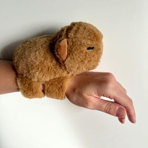Puede incluir: Una pulsera de felpa marr&oacute;n con forma de carpincho. El carpincho tiene una textura suave, orejas peque&ntilde;as y un ojo negro cosido. La pulsera se lleva en la mu&ntilde;eca.