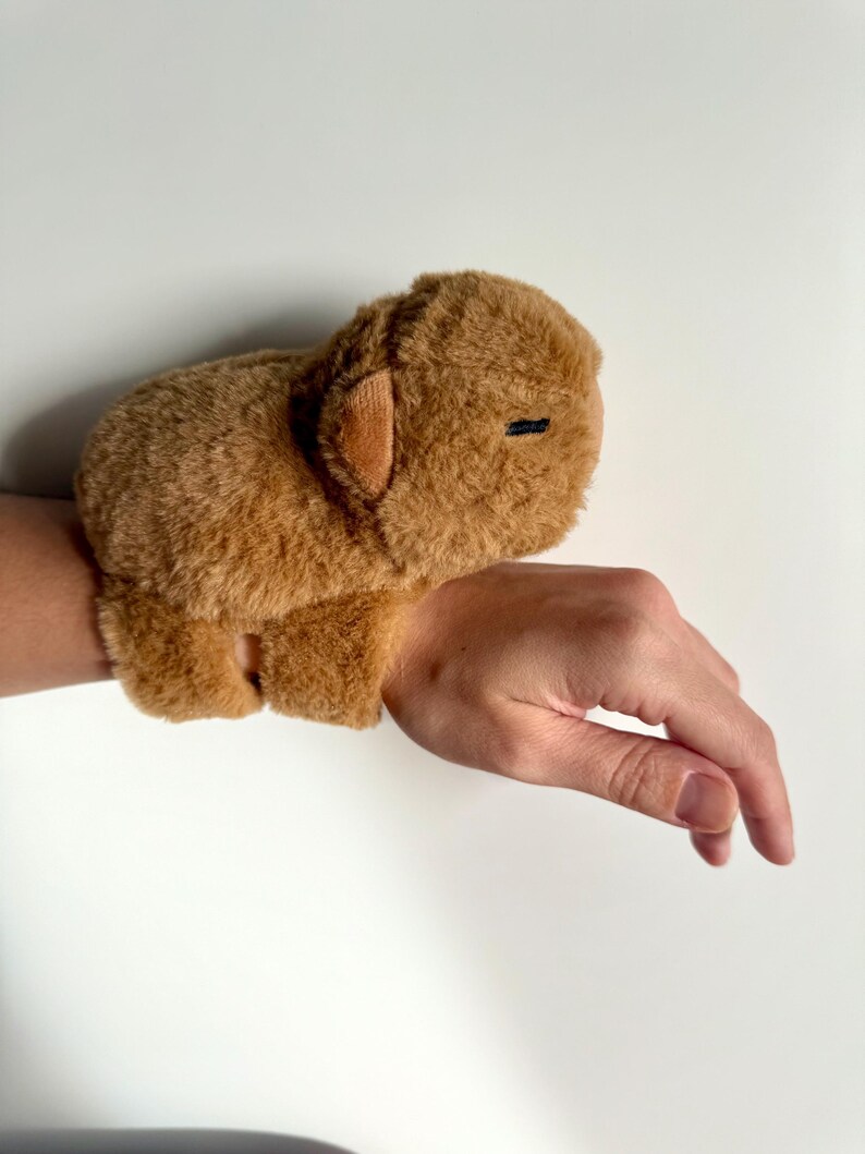 Puede incluir: Una pulsera de felpa marr&oacute;n con forma de carpincho. El carpincho tiene una textura suave, orejas peque&ntilde;as y un ojo negro cosido. La pulsera se lleva en la mu&ntilde;eca.