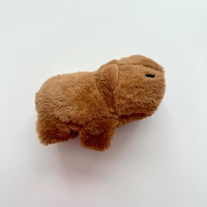 Puede incluir: Un juguete de peluche de carpincho marr&oacute;n. El peluche tiene una textura suave y peluda y peque&ntilde;os ojos negros. El carpincho est&aacute; tumbado sobre una superficie blanca.