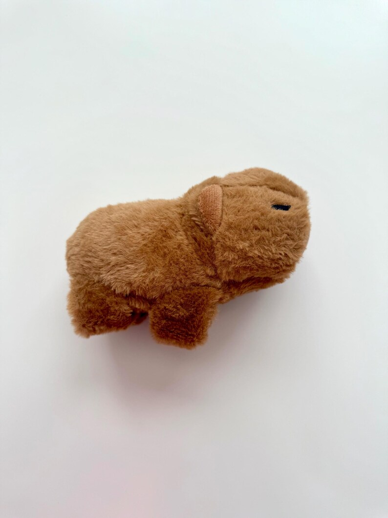 Puede incluir: Un juguete de peluche de carpincho marr&oacute;n. El peluche tiene una textura suave y peluda y peque&ntilde;os ojos negros. El carpincho est&aacute; tumbado sobre una superficie blanca.