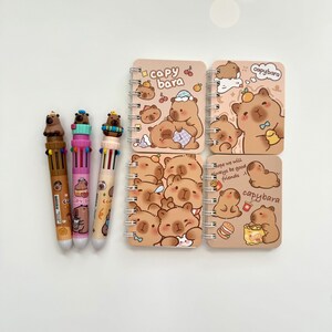 Puede incluir: Una colecci&oacute;n de art&iacute;culos de papeler&iacute;a con tem&aacute;tica de carpincho. La imagen incluye cuatro cuadernos peque&ntilde;os con espiral con ilustraciones y texto de carpinchos, junto con tres bol&iacute;grafos multicolores. Los cuadernos tienen una cubierta marr&oacute;n claro.