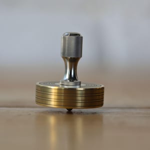 May include: A brass and silver-colored spinning top. The top has a cylindrical body with concentric grooves and a rounded top. The top is designed to spin on a pointed tip. The background is a blurred wooden surface.