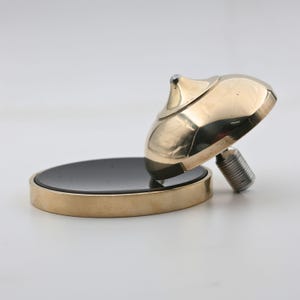 May include: A brass bottle stopper with a black circular base. The stopper has a rounded top and a threaded metal stem. The base is a dark, reflective material, and the brass has a polished finish.