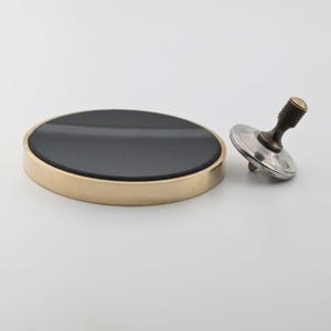 May include: A brass-rimmed, black-topped coaster and a metal spinning top with a brass stem and silver-colored base. The coaster is round and the top is tilted, suggesting motion. The items are set against a white background.