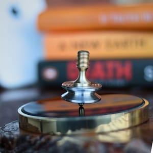 May include: A brass and silver metal spinning top with a grooved top and a rounded base. The top is spinning on a circular, gold-rimmed base. The background includes blurred books.