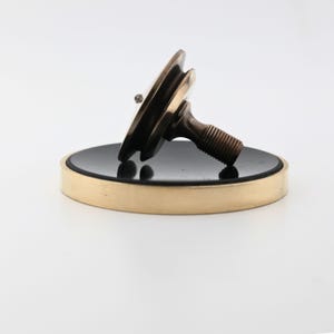 May include: A bronze-colored, decorative sink drain stopper. The stopper has a circular, black center with a gold-colored rim. The stopper has a threaded stem and a small, decorative element on top.