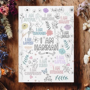 Op de afbeelding: Een wit notitieboek met bloemen- en vlinderillustraties en de tekst "I AM HANNAH". Het notitieboek bevat ook de zinnen "I AM JOYFUL", "I AM SINCERE", "I AM SMART", "I AM ENOUGH", "I AM LOVED", "I AM BOLD", "I AM KIND", "I AM LOYAL", "I AM CONFIDENT" en "I AM STRONG".