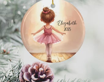 May include: Round ceramic ornament featuring a watercolor illustration of a young ballerina in a pink tutu. The ornament has the name "Elizabeth" and the year "2025" in elegant script. A gold string is attached for hanging. The background is a soft, warm tone.