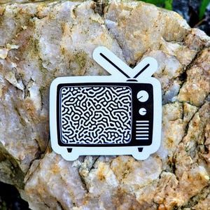 May include: A black and white sticker of a retro television set. The TV has an antenna and a screen filled with a black and white squiggly pattern. The sticker is placed on a light-colored rock with green foliage in the background.