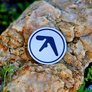 May include: A white circular sticker with a dark blue logo, featuring a stylized, abstract shape. The sticker is set against a textured, light brown rock with green foliage in the background.