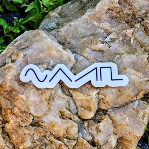 May include: A white sticker with a black line design. The design features a wavy line, two upward-pointing triangles, and a square. The sticker is placed on a light brown and tan rock with green foliage in the background.