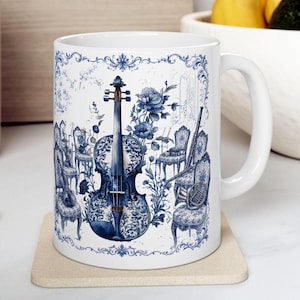 May include: White ceramic mug with a blue and white design depicting a violin and an orchestra. The illustration includes floral patterns and decorative chairs, similar to traditional porcelain. The mug has a curved handle.
