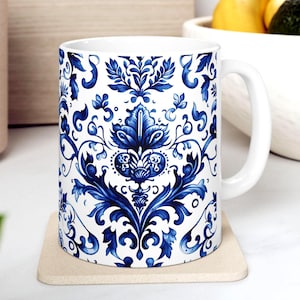 May include: White ceramic mug with a blue floral pattern. The design features intricate blue botanical elements on a white background. The mug has a curved handle and sits on a beige coaster.