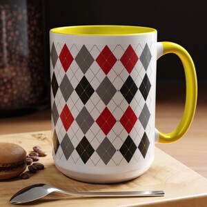 May include: A white ceramic mug with a yellow interior and handle. The mug features a repeating argyle pattern in red, black, and gray diamonds. A silver spoon and a macaron are on a wooden surface.