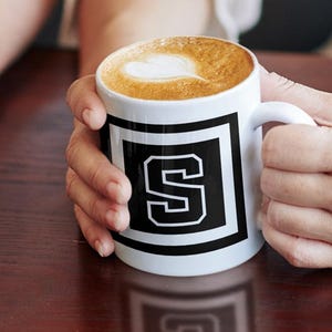 Op de afbeelding: Twee witte koffiemokken met zwarte vierkante randen en grote witte letters, een met een "T" en de andere met een "S". Elke mok is gevuld met koffie en afgewerkt met een hartvormig ontwerp. De mokken staan op een donkerbruin oppervlak.