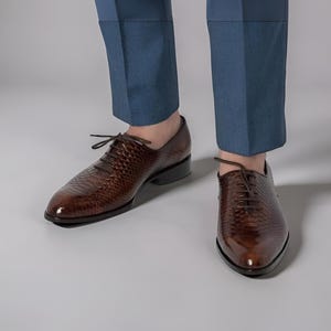 May include: A pair of dark brown lace-up dress shoes with a textured snakeskin-like pattern and a polished finish. The shoes feature a pointed toe, thin dark brown laces, and a low heel, worn with blue trousers that end just above the ankle.
