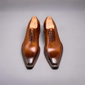 Op de afbeelding: Een paar bruine leren herenschoenen met een glanzende afwerking. De schoenen hebben een klassiek Oxford-ontwerp met een puntige neus en vetersluiting. De zolen zijn donkerbruin, tegen een grijze achtergrond.