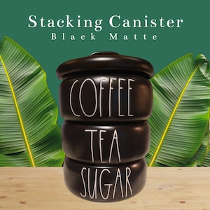 May include: A matte black stacking canister with a rounded lid. The canister features three distinct stacked sections with white text printed on each, reading COFFEE, TEA, and SUGAR in a tall, thin, handwritten-style font. The surface is smooth.