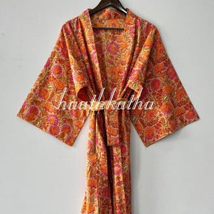 May include: A floral print robe in shades of orange, pink, and beige. The robe features a tie closure and wide sleeves. The text "haathkatha" is printed on the robe. This garment is suitable for lounging or as a light cover-up.