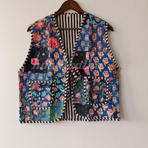 May include: A quilted vest with a patchwork design featuring floral and geometric patterns in shades of blue, red, and green. The vest has a striped interior and is hanging on a wooden hanger.