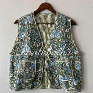 May include: Olive green quilted vest with a floral pattern in blue and white. The vest has two pockets and a striped lining. The vest is hanging on a wooden hanger.