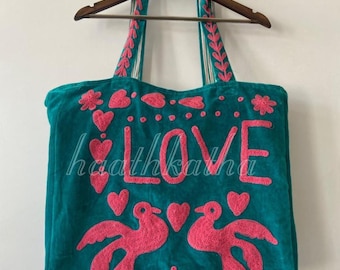 May include: Teal tote bag with pink embroidered details. The word "LOVE" is embroidered in large pink letters. Pink hearts, flowers, and birds decorate the bag. The handles and top trim are also embroidered.