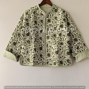 Puede incluir: Chaqueta acolchada blanca y verde oliva con estampado floral. La chaqueta tiene cuello mandarín, dos bolsillos delanteros y cierre de cremallera. Las mangas son de tres cuartos y los puños tienen un diseño a rayas. La chaqueta está colgada en una percha de madera.