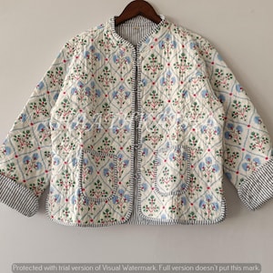 Puede incluir: Chaqueta acolchada color crema con un estampado floral en azul, rojo y verde. La chaqueta tiene cuello mandarín, dos bolsillos y puños a rayas. La chaqueta se muestra en una percha de madera.