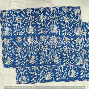 May include: Two blue placemats with a white floral pattern. The placemats are rectangular with a stitched border. The floral design includes stylized flowers and leaves. The text "haathkatha" is visible on the placemats.