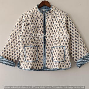 May include: Cream-colored quilted jacket with a floral pattern in blue and brown. The jacket has a mandarin collar, two front pockets, and blue striped cuffs. The jacket is hanging on a wooden hanger.