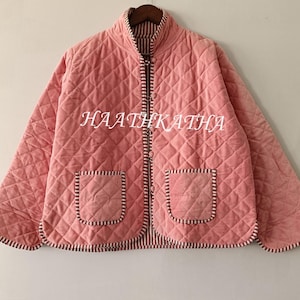 Op de afbeelding: Een gewatteerde roze jas met een hoge kraag, twee voorzakken en een ritssluiting. De jas heeft een ruitpatroon en is afgewerkt met een gestreepte stof. De tekst "HAATHKATHA" is op de voorkant gedrukt.