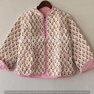 Puede incluir: Chaqueta acolchada con estampado floral rosa y verde sobre fondo blanco. La chaqueta tiene cierre de cremallera, dos bolsillos y ribete a rayas rosas. Cuelga de una percha de madera.