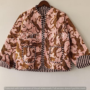 Pode incluir: Casaco reversível com estampa de leopardo marrom em fundo rosa. O casaco tem acabamento listrado em preto e branco nos punhos, bolsos e gola. Está pendurado em um cabide de madeira.