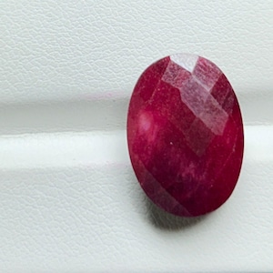 May include: A faceted, oval-shaped ruby gemstone. The ruby is a deep, rich red color with a polished surface. The facets create a play of light across the surface of the stone. The gemstone is set against a white background.