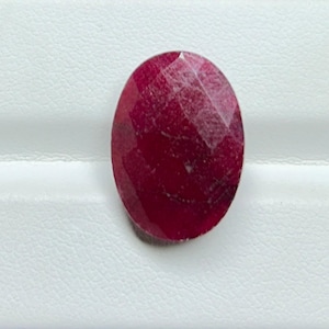 May include: A faceted, oval-shaped ruby gemstone. The gem is a deep, rich red color, with visible internal inclusions and a polished surface. The ruby is set against a white background.