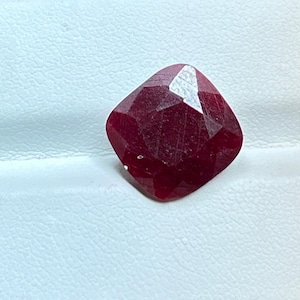 Könnte beinhalten: Ein facettierter Rubin-Edelstein im Kissenschliff. Der Edelstein hat eine tiefe, satte rote Farbe mit sichtbaren inneren Einschlüssen. Der Schliff des Steins erzeugt mehrere reflektierende Oberflächen, was seinen Glanz verstärkt. Der Rubin liegt auf weißem Hintergrund.