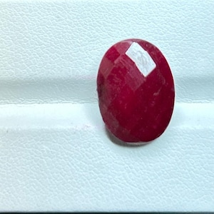 Könnte beinhalten: Ein facettierter, ovaler Rubin-Edelstein. Der Edelstein hat eine tiefe, satte rote Farbe mit einer polierten Oberfläche. Die Facetten fangen das Licht ein und erzeugen Glanzlichter und Schatten. Der Rubin liegt vor einem weißen Hintergrund.