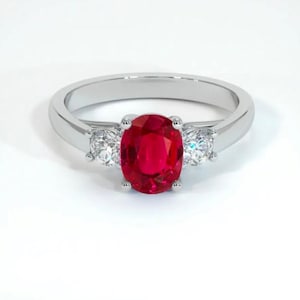 Könnte beinhalten: Ein silberner Ring mit einem großen, oval geschliffenen Rubin als Mittelstein, flankiert von zwei kleineren, runden, klaren Edelsteinen. Der Ring hat eine polierte, reflektierende Oberfläche, vor einem weißen Hintergrund. Ein elegantes Schmuckstück.