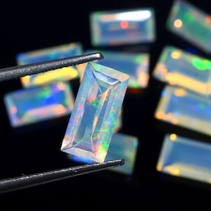 Puede incluir: Colección de gemas de ópalo rectangulares con destellos de color iridiscente. Una gema es sostenida por unas pinzas, mostrando sus facetas pulidas y su juego de color interno. Los ópalos se presentan sobre un fondo oscuro.