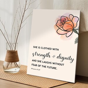 Peut inclure: Impression sur toile avec l'inscription « SHE IS CLOTHED WITH strength & dignity AND SHE LAUGHS WITHOUT FEAR OF THE FUTURE. » Une rose à l'aquarelle pêche et rose est en haut à droite. Un vase en verre avec des branches est à gauche.