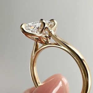May include: A gold ring with a large, clear, pear-shaped gemstone. The ring's band is a smooth, polished gold, and the gemstone is held in place by delicate prongs. The ring is shown against a soft, neutral background.
