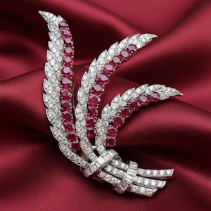 May include: A silver-toned brooch in a floral design, set with red gemstones and clear crystals. The brooch features three stylized feather-like elements, each adorned with a row of red stones and outlined with smaller clear crystals. The base of the brooch has a ribbon-like design.